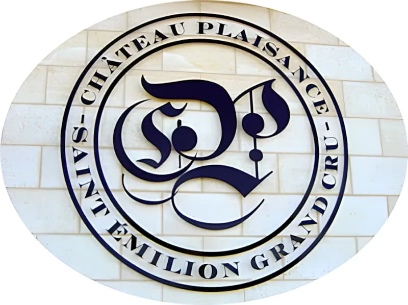 安装在白色石墙上的圆形金属标识，以哥特式字体装饰「CP」首字母，边框有「Château Plaisance」和「Saint-Émilion Grand Cru」文字。