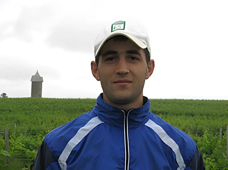 Un jeune homme souriant portant une veste de survêtement bleue et blanche et une casquette, avec un château d'eau visible dans l'arrière-plan de campagne verte.