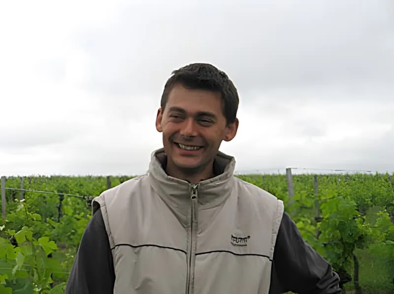 Un jeune homme joyeux en veste de couleur claire debout devant des rangs de vignoble verts luxuriants, souriant chaleureusement à la caméra.