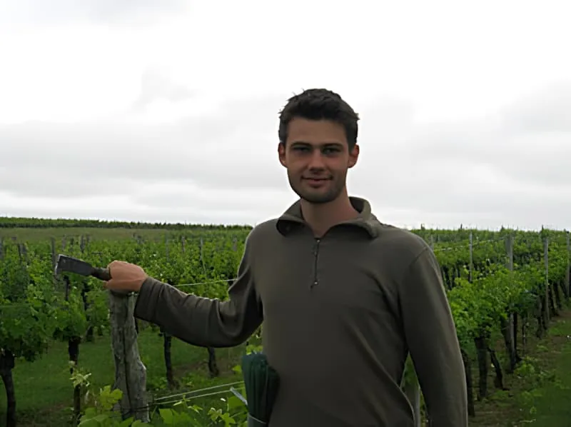 Un jeune homme en vêtements sombres tenant un outil de vignoble tout en se tenant parmi les vignes vertes, montrant le travail pratique du vignoble.